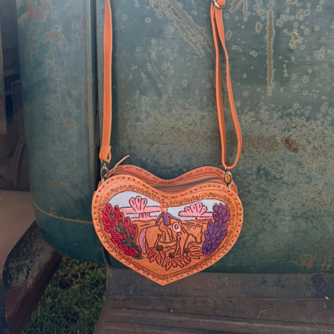 A heart-shaped leather crossbody bag with a western-themed cowboy and cowgirl illustration, hanging in front of a rustic green metal barrel.