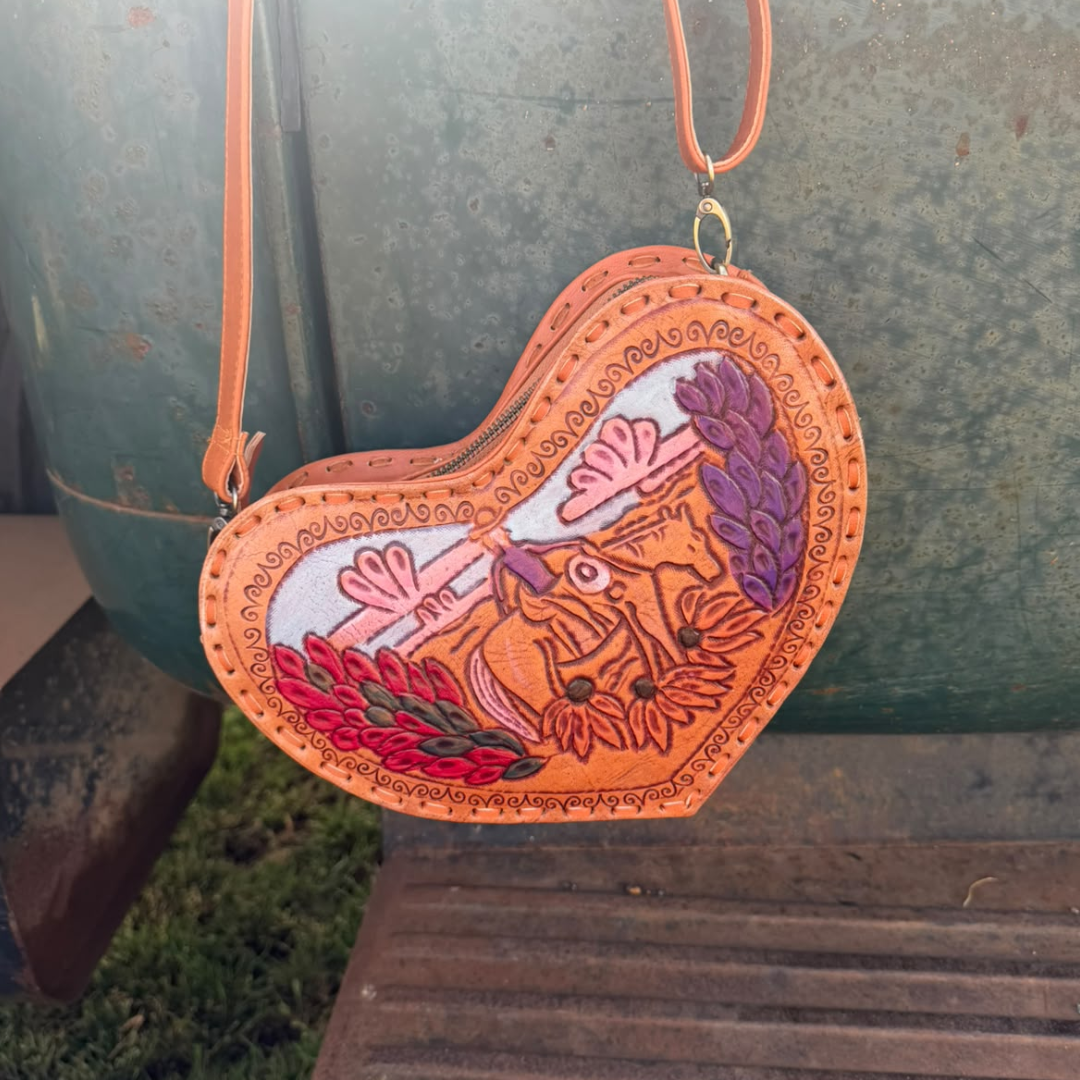 A heart-shaped leather crossbody bag with a colorful embroidered design of a horse, flowers, and leaves, shown hanging outdoors against a rustic green metal container.