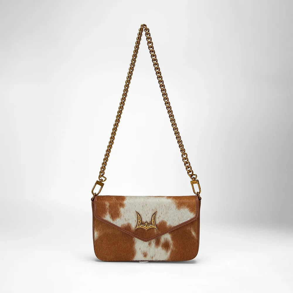 House of Hide genuine leather brown and white cowhide shoulder bag with a gold-tone chain strap and gold bat-shaped logo on the front flap against a light gray studio background