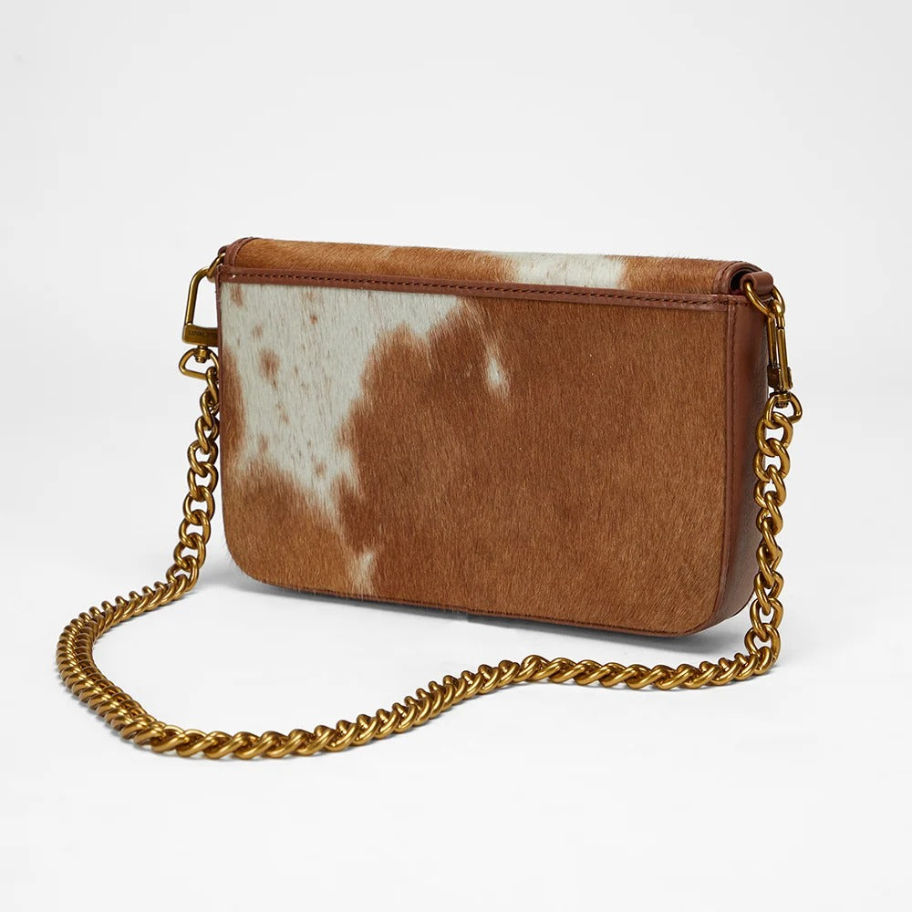 Close-up of a House of Hide genuine leather brown and white cowhide shoulder bag featuring a gold-tone chain strap and bat-shaped logo on the front flap against a light gray studio background