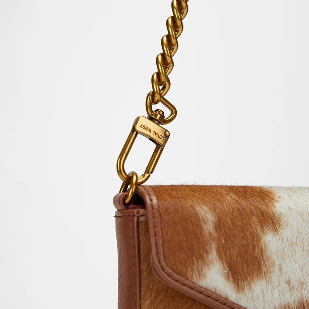 Close-up of a House of Hide genuine leather brown and white cowhide handbag featuring a gold chain strap on a light gray studio background
