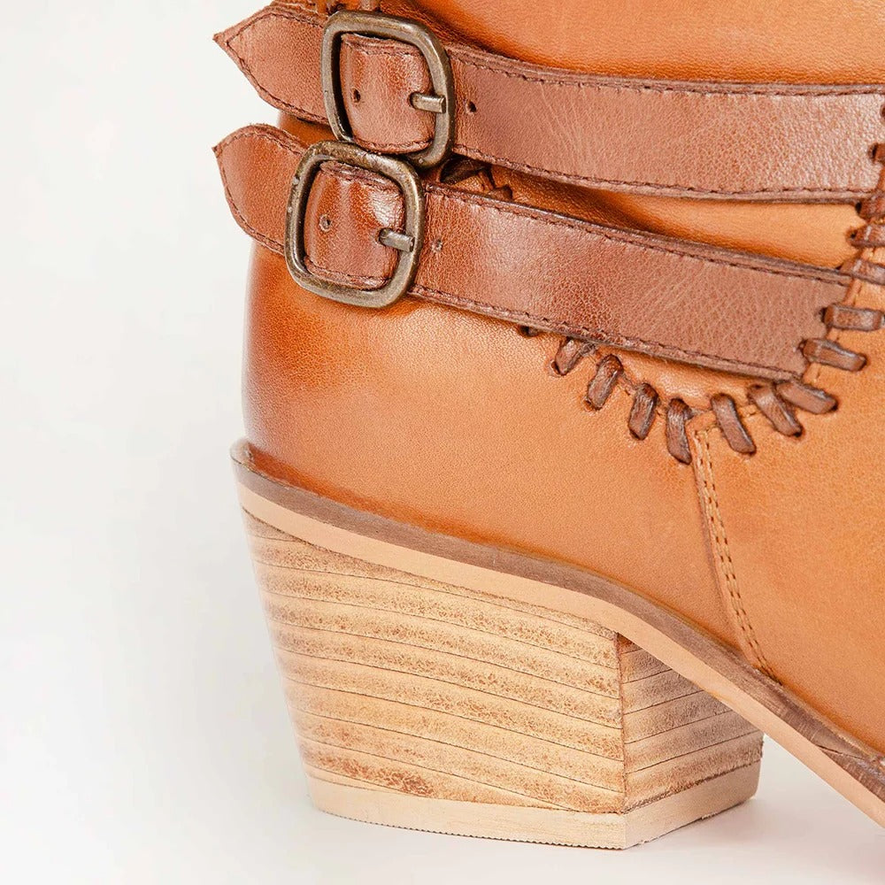 Close-up of a tan leather ankle boot with double buckle straps, decorative stitching, and a stacked wood-look block heel on a white background.
