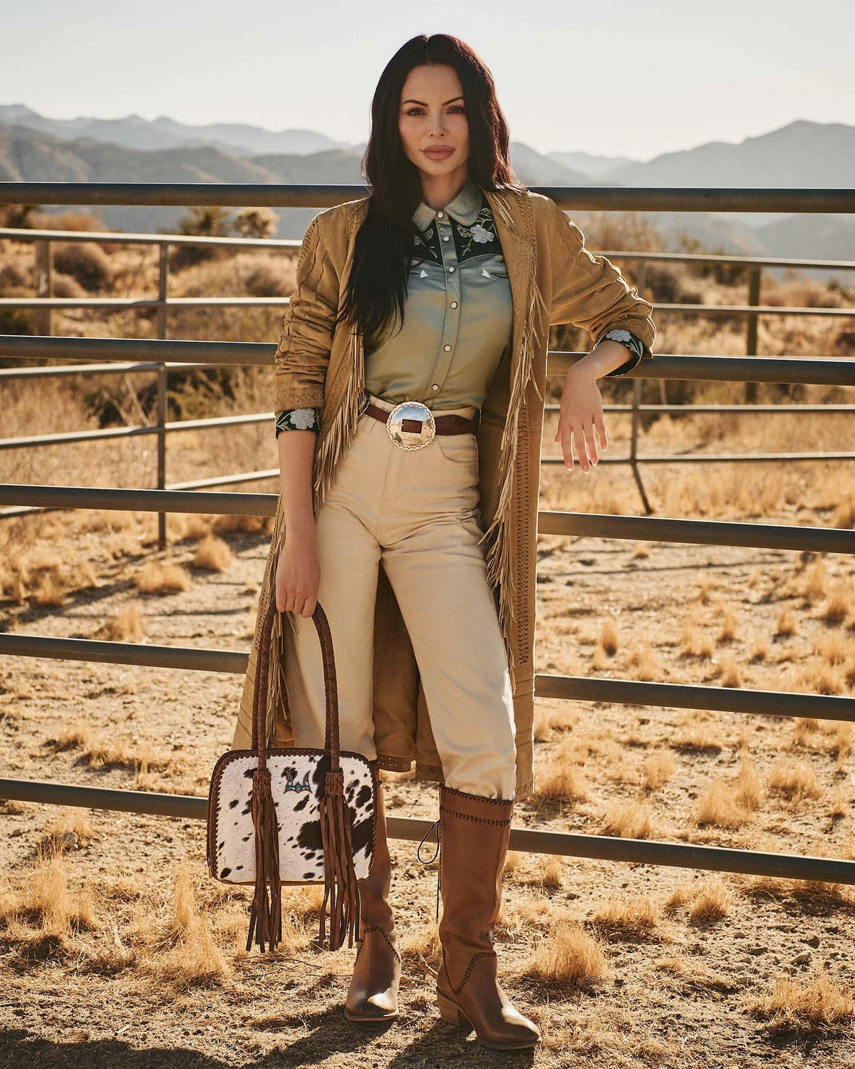 Woman in western attire standing in a desert landscape with mountains in the background