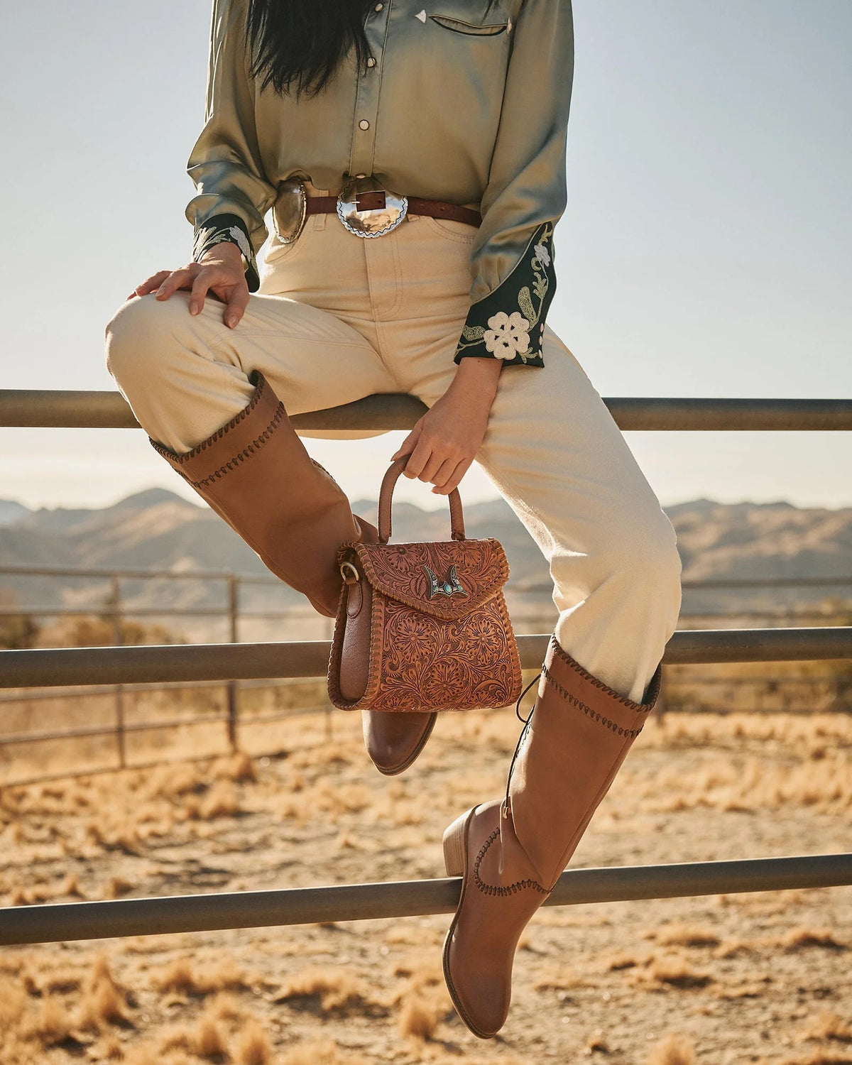 Model wearing House of Hide western fashion sitting on a fence with a scenic desert landscape in the background