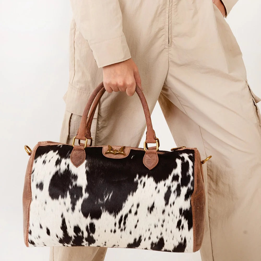 A person in beige pants holds a large duffel bag with a black and white cowhide pattern and brown leather handles against a plain white background.
