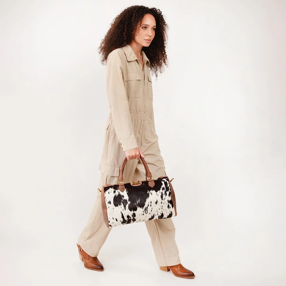 A model with curly dark hair is shown in profile wearing a beige utility jumpsuit and brown leather boots, holding a black and white cow-print handbag with brown leather handles, set against a plain white studio background.