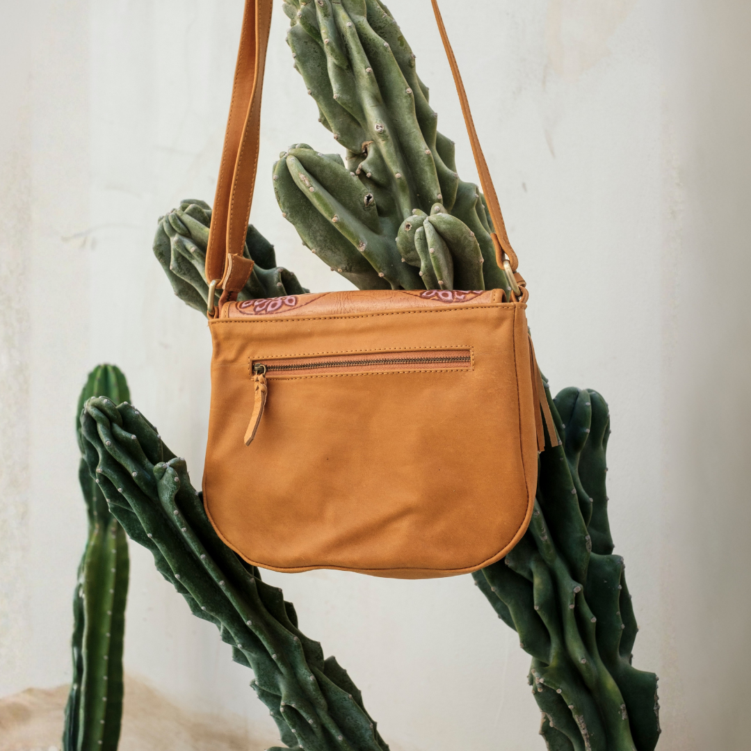 Magnolia Crossbody Bag from House of Hide in tan leather with front zip pocket and embroidered detailing being displayed hanging against white wall with green cactus