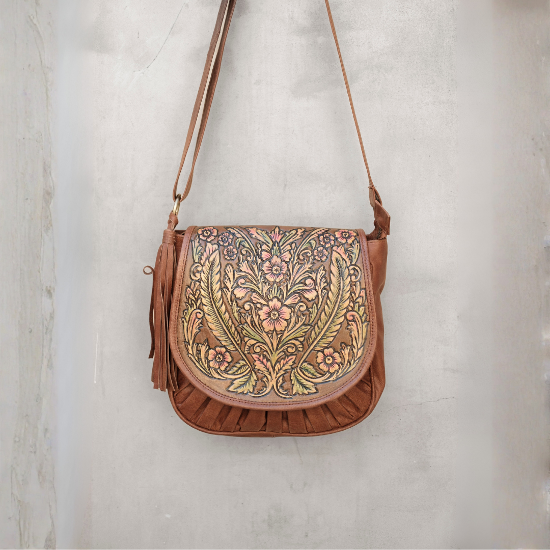 Meadow Crossbody Bag from House of Hide in brown leather with intricate floral and leaf pattern on front flap being displayed hanging against textured light gray wall