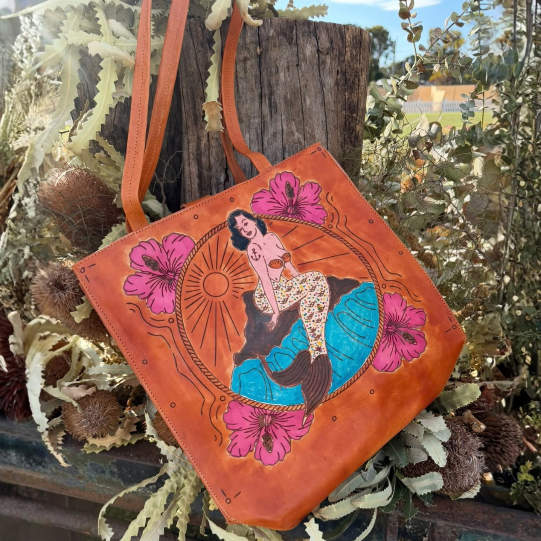 An orange-brown leather tote bag with a hand-painted mermaid illustration and pink flowers, displayed outdoors against a rustic wooden post and dried foliage.