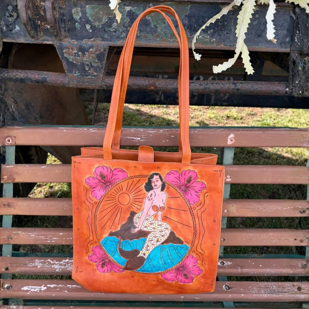 An orange leather tote bag with a hand-painted mermaid illustration and pink hibiscus flowers, resting on a weathered wooden bench.