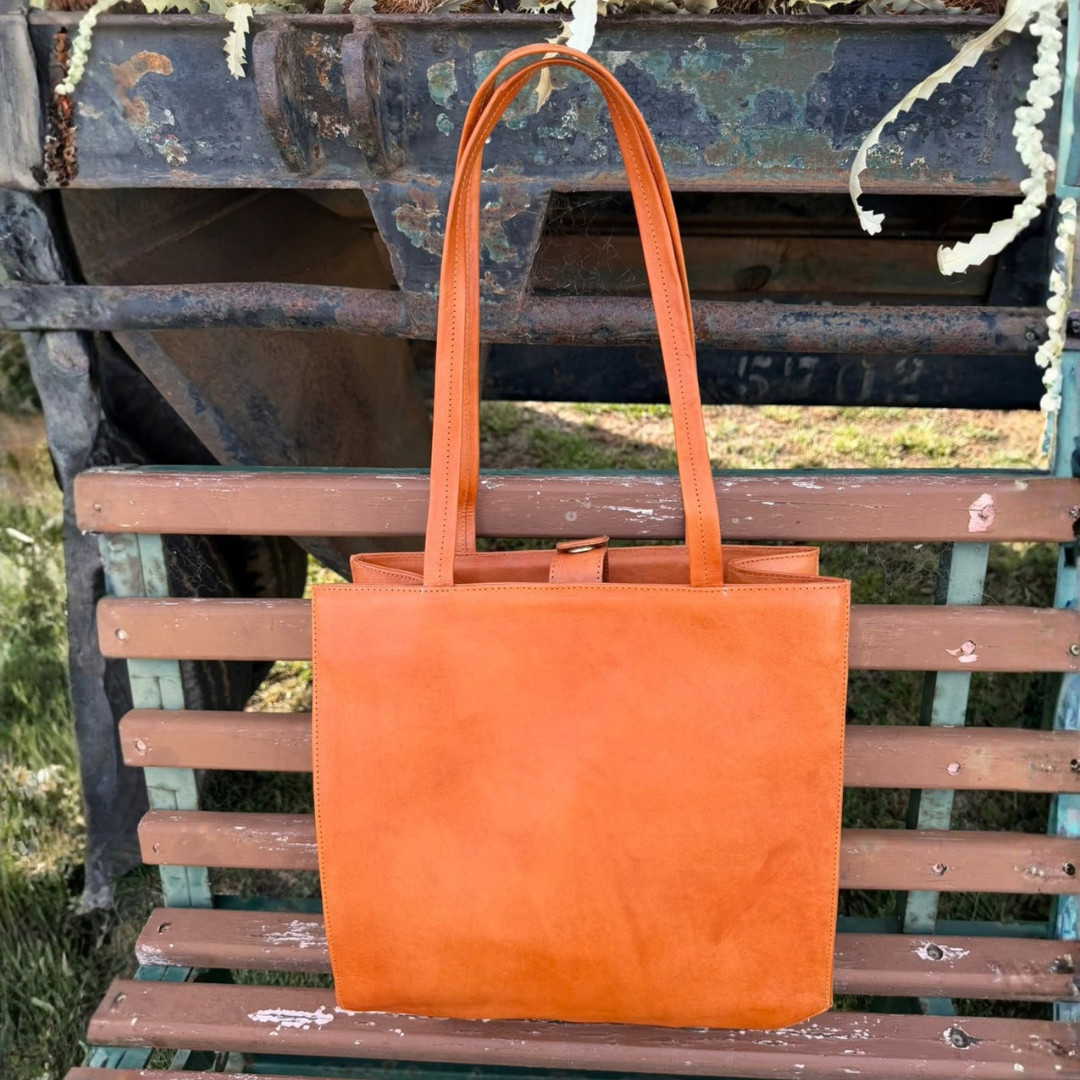 An orange leather tote bag with two shoulder straps, displayed on a weathered wooden bench outdoors.