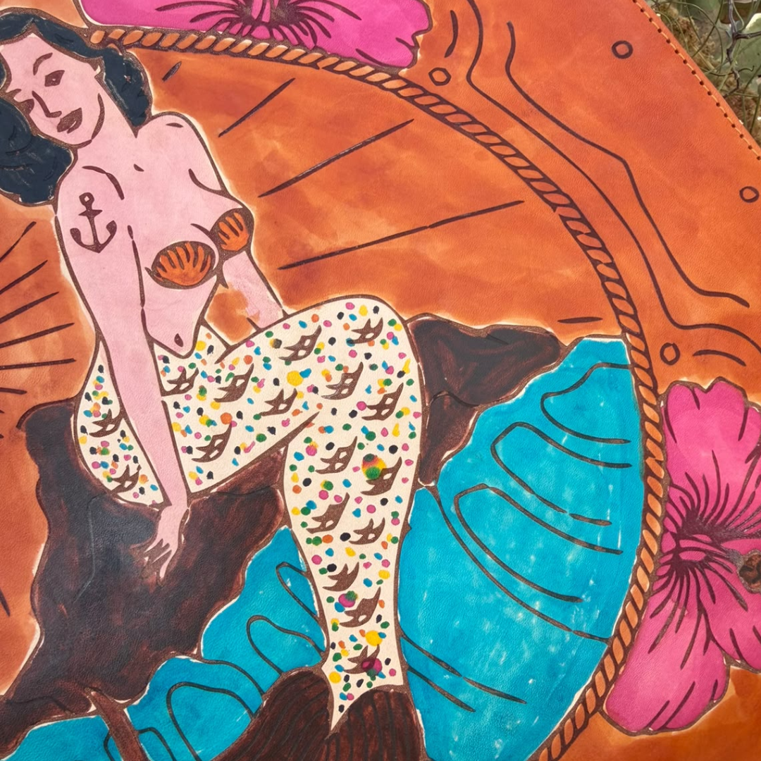 A close-up of a colorful painted artwork showing a stylized mermaid with a pink skin tone, patterned tail, orange background, and bright blue water, with part of a person's head and body also visible in the scene.