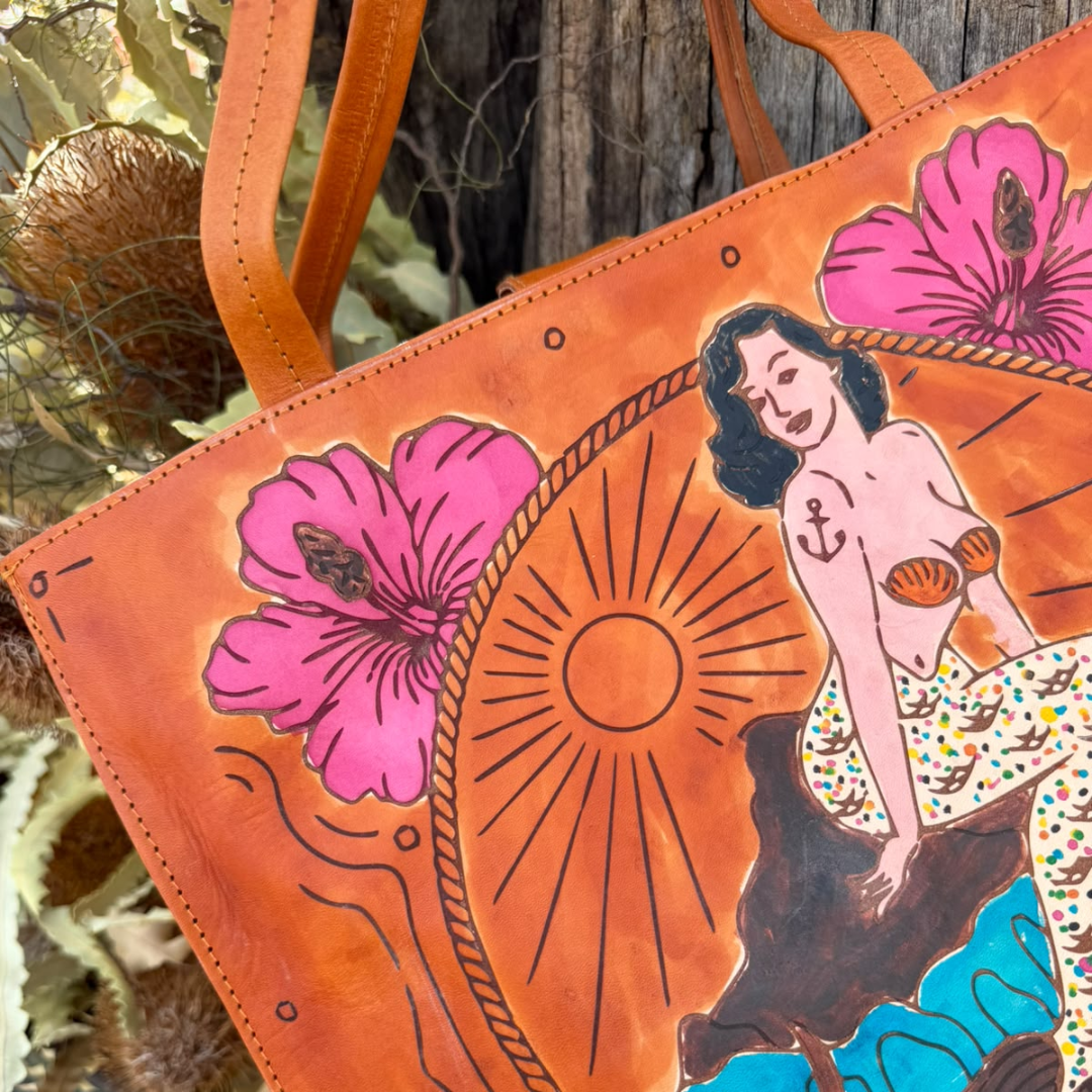 A close-up of a tan leather tote bag with a hand-painted retro pin-up illustration and pink hibiscus flowers, set against a rustic wooden and natural background.