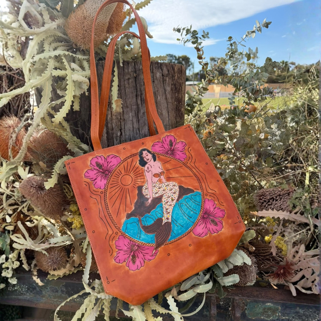 A tan leather tote bag with a hand-painted mermaid illustration, displayed outdoors among dried plants and a rustic wooden post.