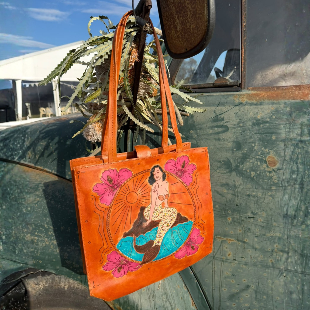 An orange leather tote bag with a colorful mermaid illustration and pink hibiscus flowers, hanging from a vintage green truck outdoors.