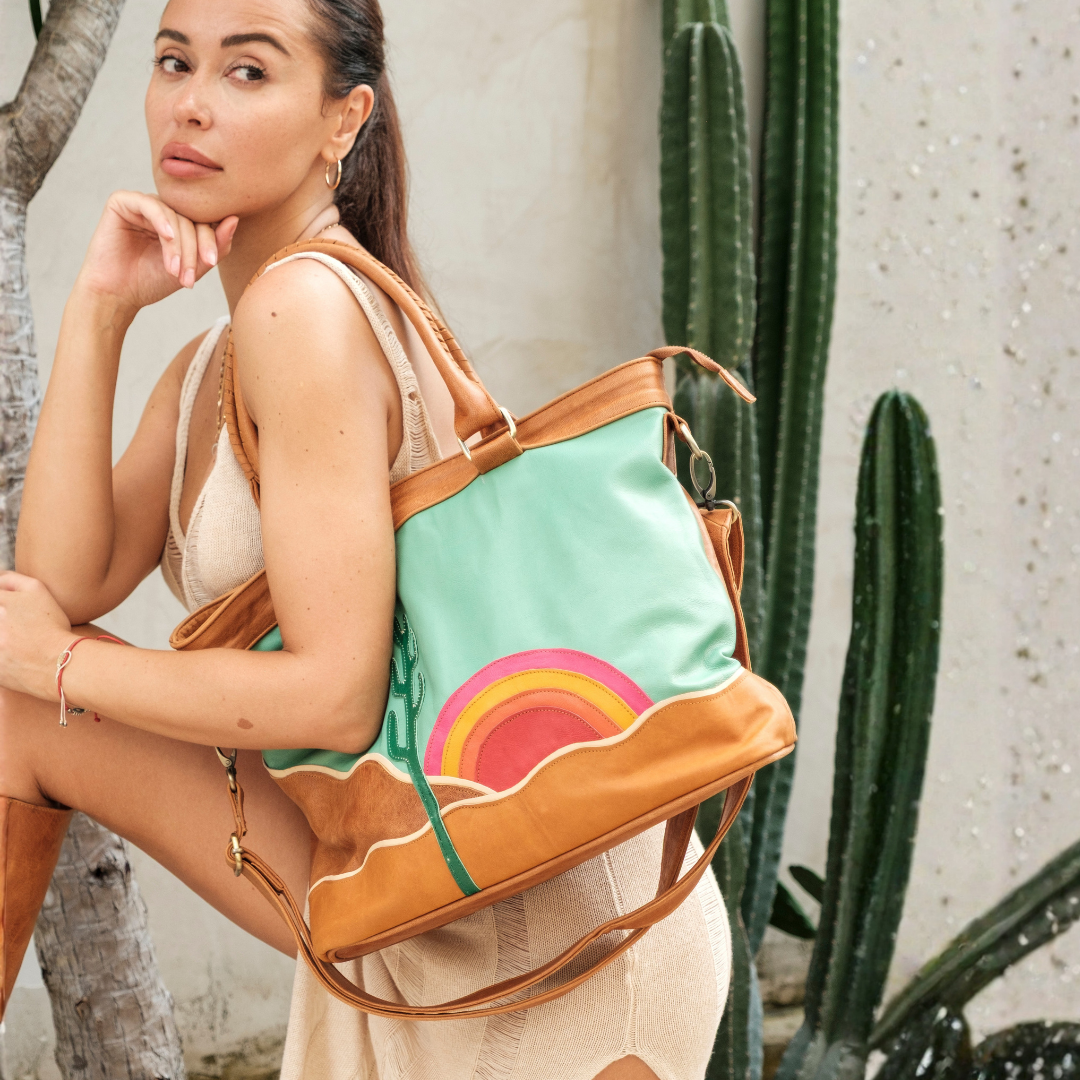 Mesa Sunset Tote Bag from House of Hide in mint green and tan leather with colorful rainbow design on front being carried by model outdoors next to cacti
