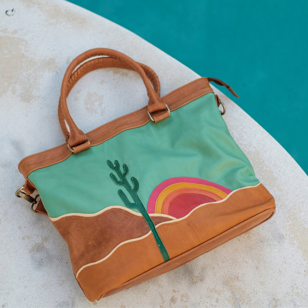 Mesa Sunset Tote Bag from House of Hide in green and brown leather with cactus and rainbow landscape design being displayed on white surface next to turquoise pool