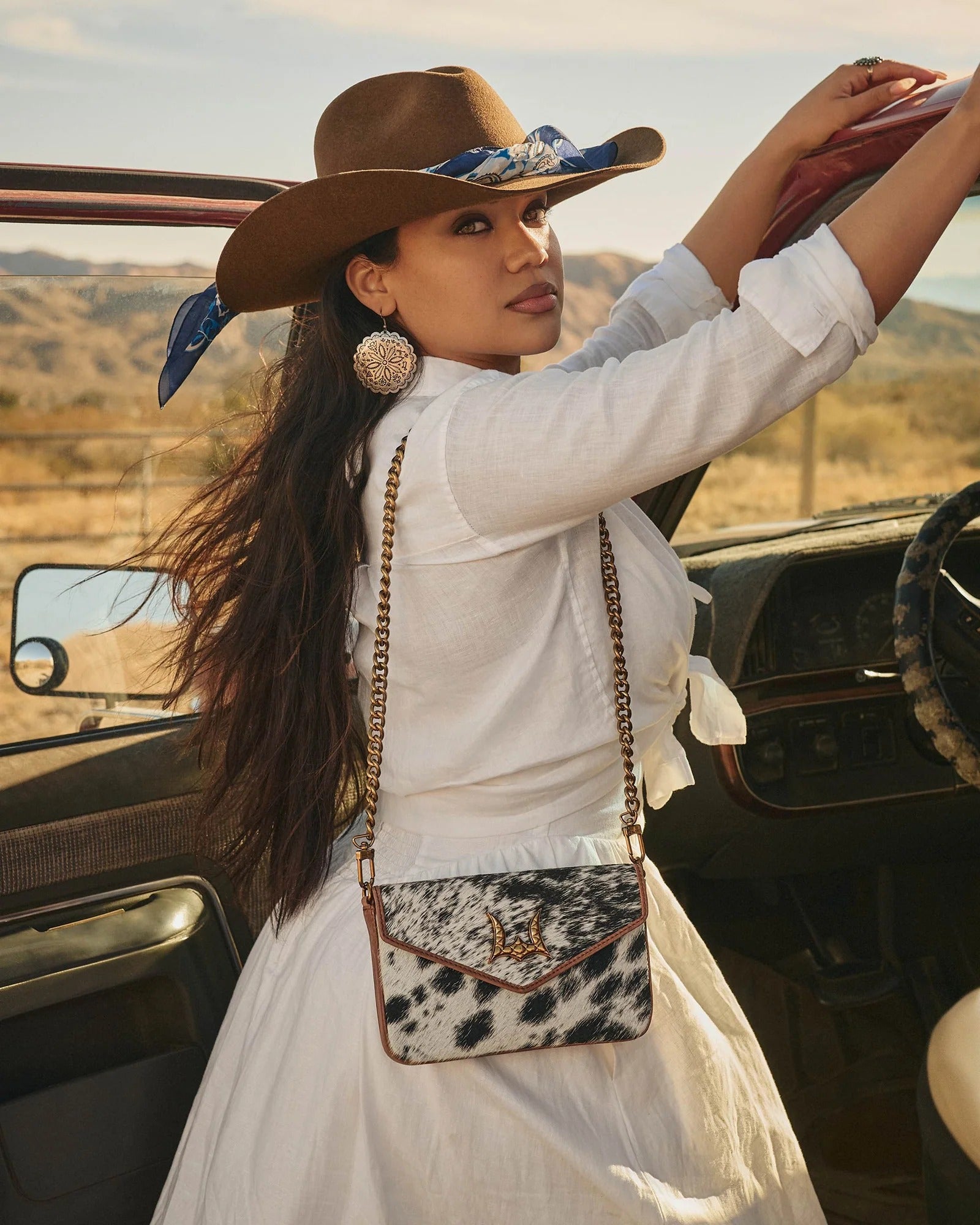 Woman in a white dress and brown hat with a leopard print purse standing next to a vehicle in a desert setting.