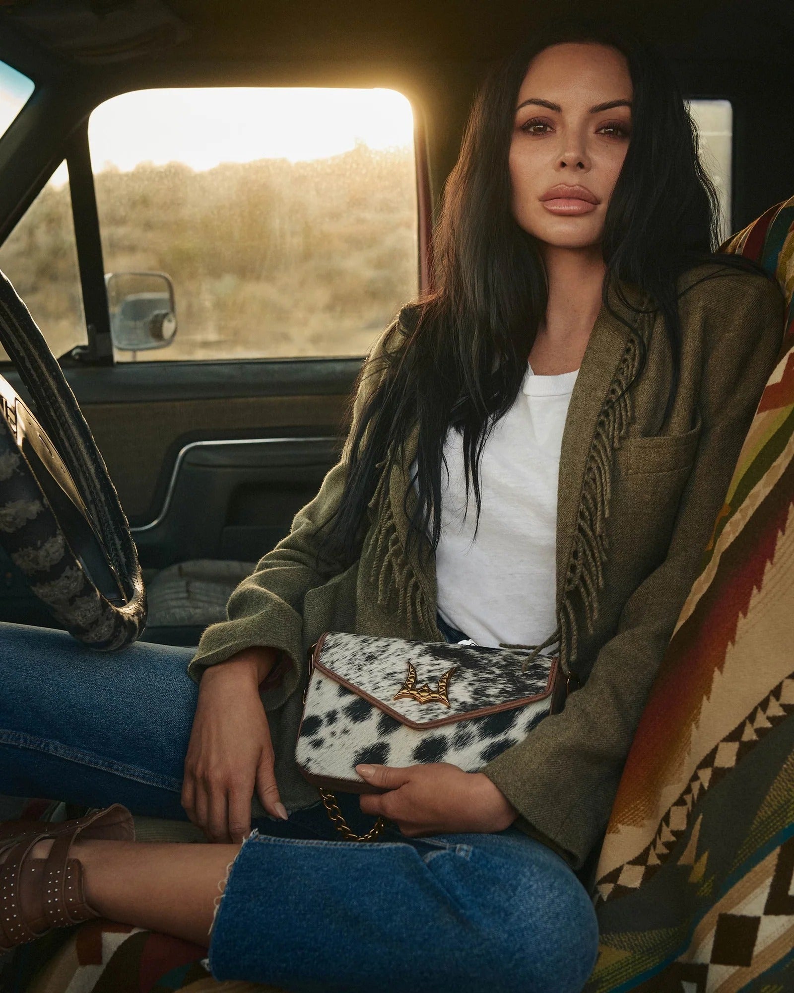 Model showcasing House of Hide western fashion while seated inside a vehicle holding a genuine leather patterned clutch with a scenic background