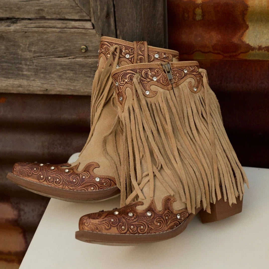 A pair of western-style ankle boots with long tan fringe, tooled brown leather detailing, and small silver rhinestone accents, displayed on a white surface against a rustic wooden and metal background.