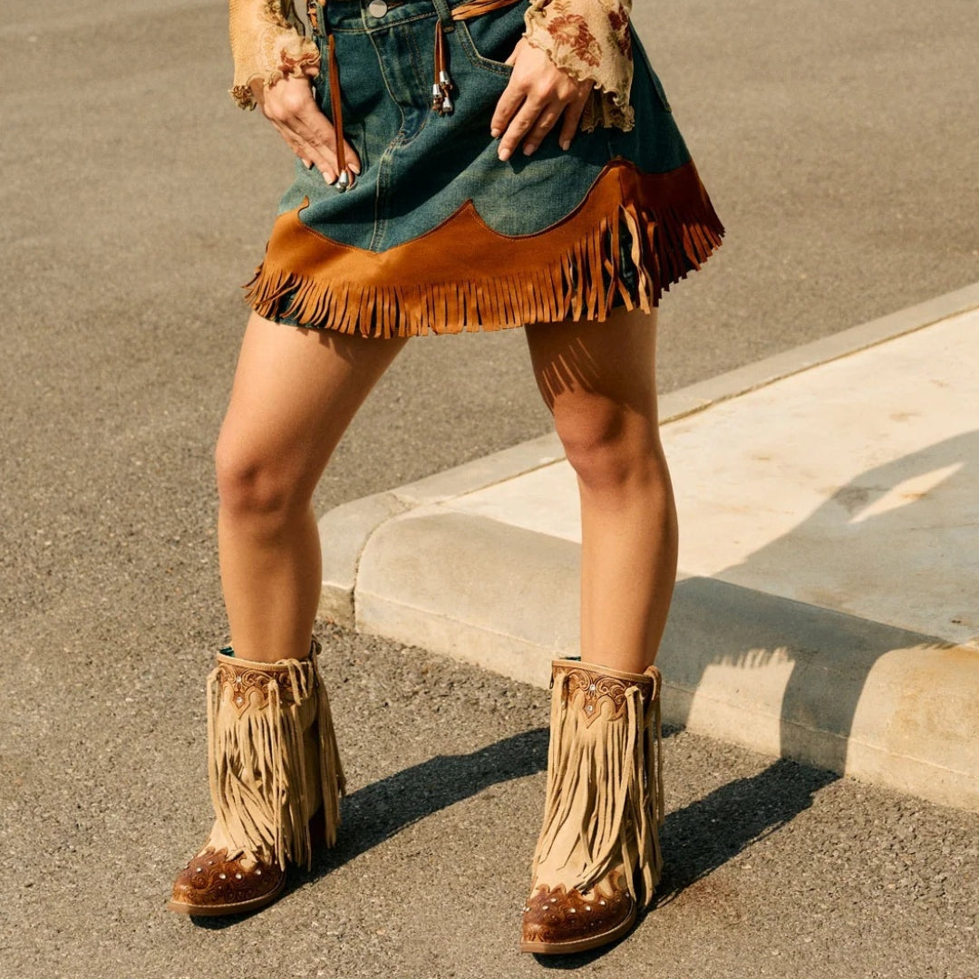 A person outdoors wearing a denim mini skirt with a brown fringe hem, a floral long-sleeve top, and tan fringe ankle boots on an asphalt street next to a curb.
