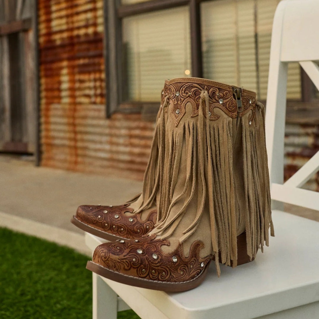 A pair of tan and brown fringed western-style boots with tooled leather detailing, displayed on a white chair in an outdoor setting.