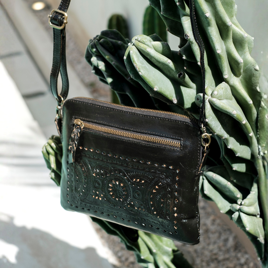 Nightshade Crossbody Bag from House of Hide in black leather with embossed geometric patterns and gold-tone hardware being displayed against green cactus outdoors