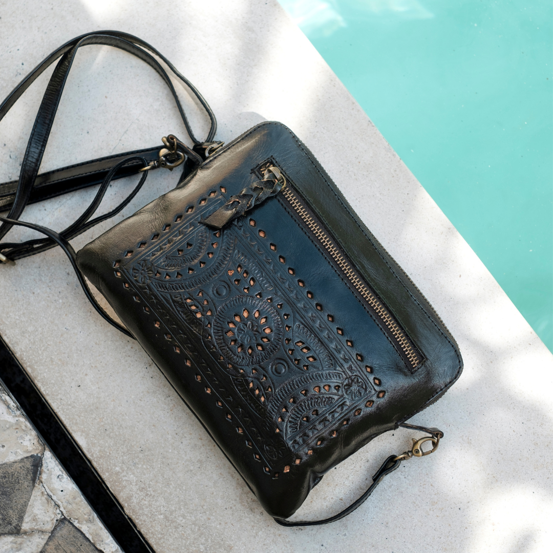 Nightshade Crossbody Bag from House of Hide in black leather with embossed floral pattern and perforated cutouts, gold-toned zipper being displayed on light stone near turquoise pool