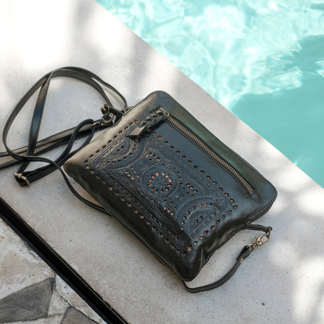 Nightshade Crossbody Bag from House of Hide in black leather with laser-cut embossed patterns and gold-tone zipper being displayed on concrete poolside deck