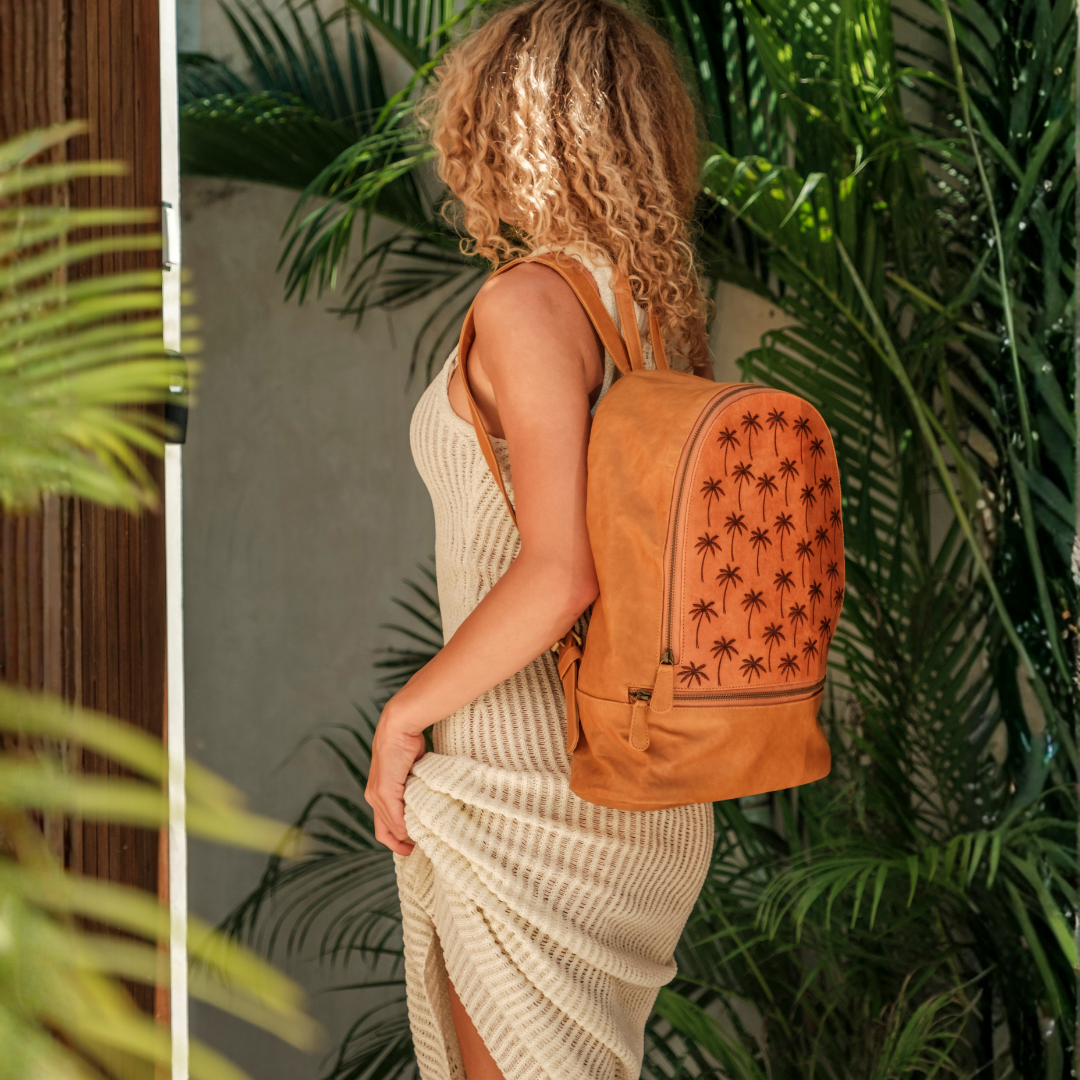 Palm Print Leather Backpack from House of Hide in tan leather with black palm tree pattern being worn from back view by model against green tropical plants background