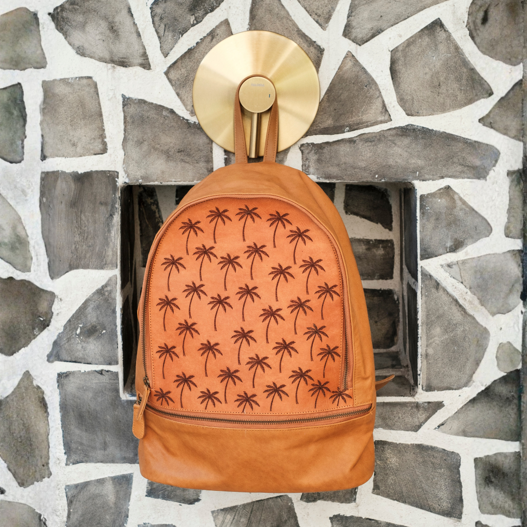 Palm Print Leather Backpack from House of Hide in orange-brown leather with embossed palm tree pattern being displayed on gold wall hook against gray stone fireplace