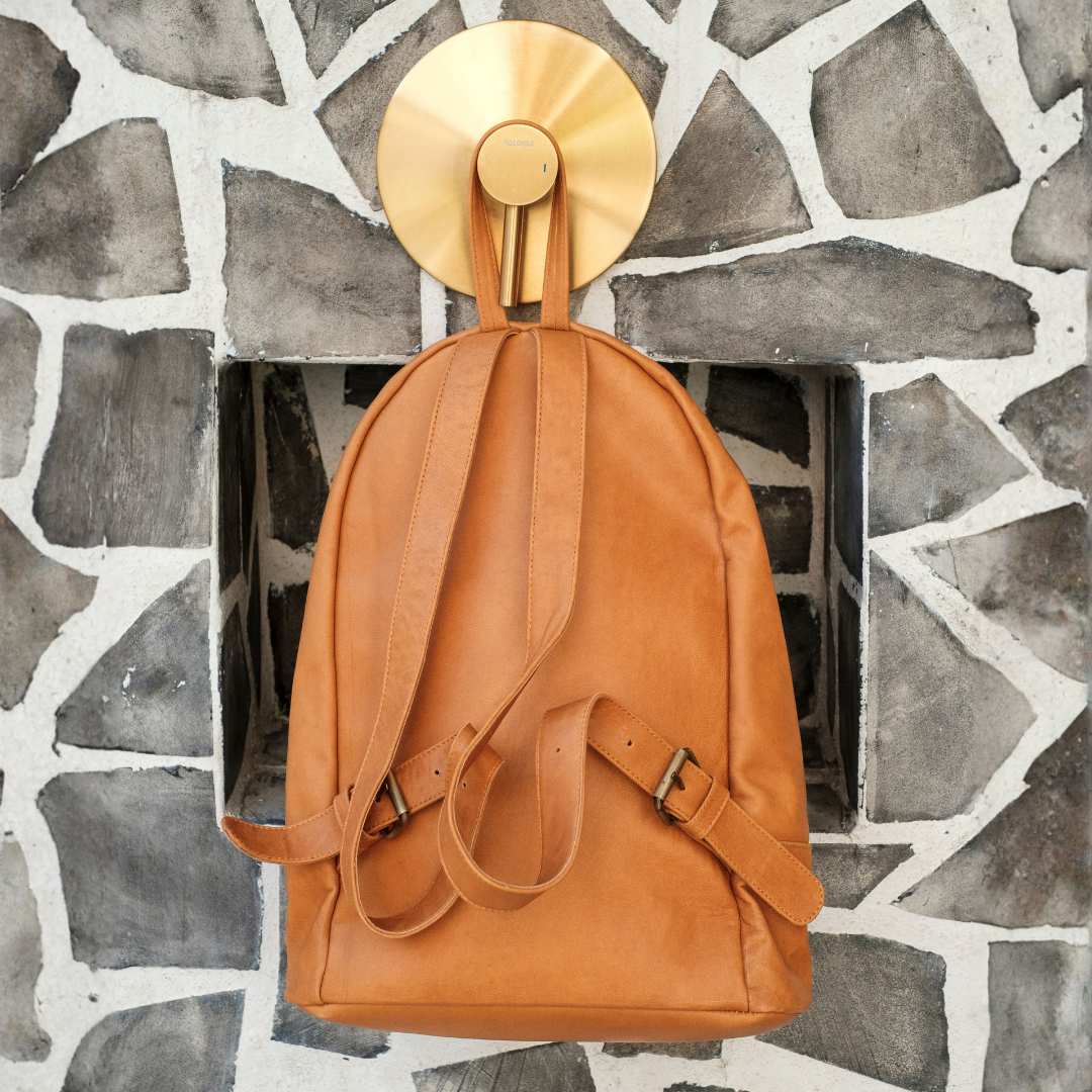 Palm Print Leather Backpack from House of Hide in tan leather being displayed hanging from round gold wall hook against gray stone wall