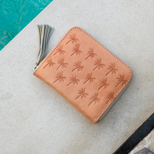 Palm Springs Zip Wallet from House of Hide in tan leather with embossed palm tree pattern and gray tassel zipper being displayed on light stone near turquoise pool