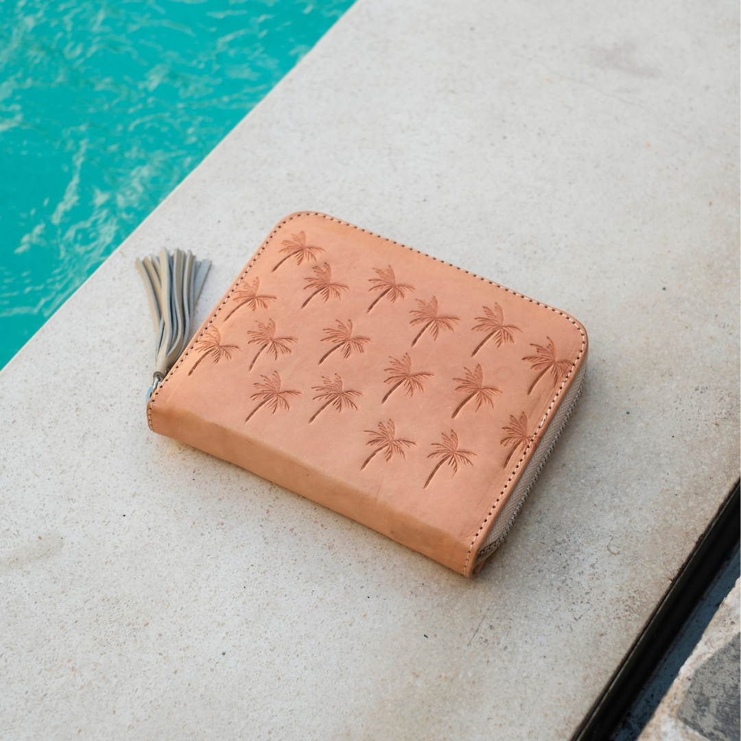 Palm Springs Zip Wallet from House of Hide in tan leather with embossed palm tree pattern and tassel zipper being displayed on light stone surface next to swimming pool