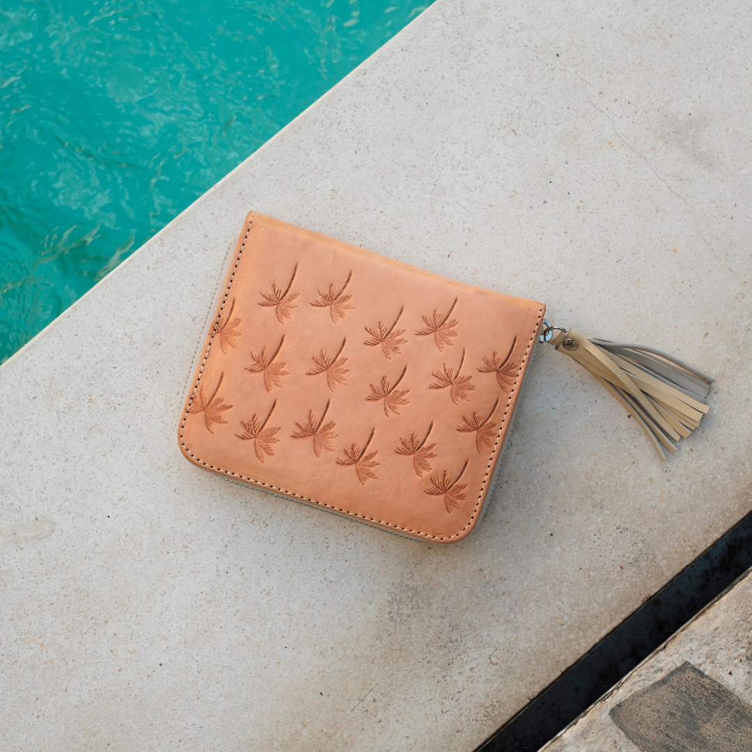 Palm Springs Zip Wallet from House of Hide in tan leather with embossed dragonfly pattern and beige tassel being displayed on light stone near turquoise pool