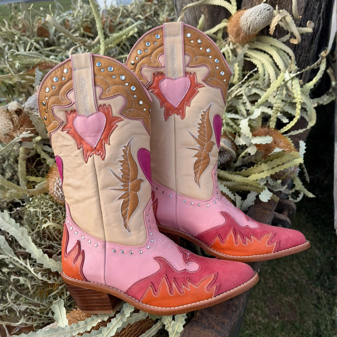 A pair of western cowboy boots with a pink, orange, and beige color-blocked design, featuring embroidered hearts, birds, and flames, displayed outdoors among dried plants.