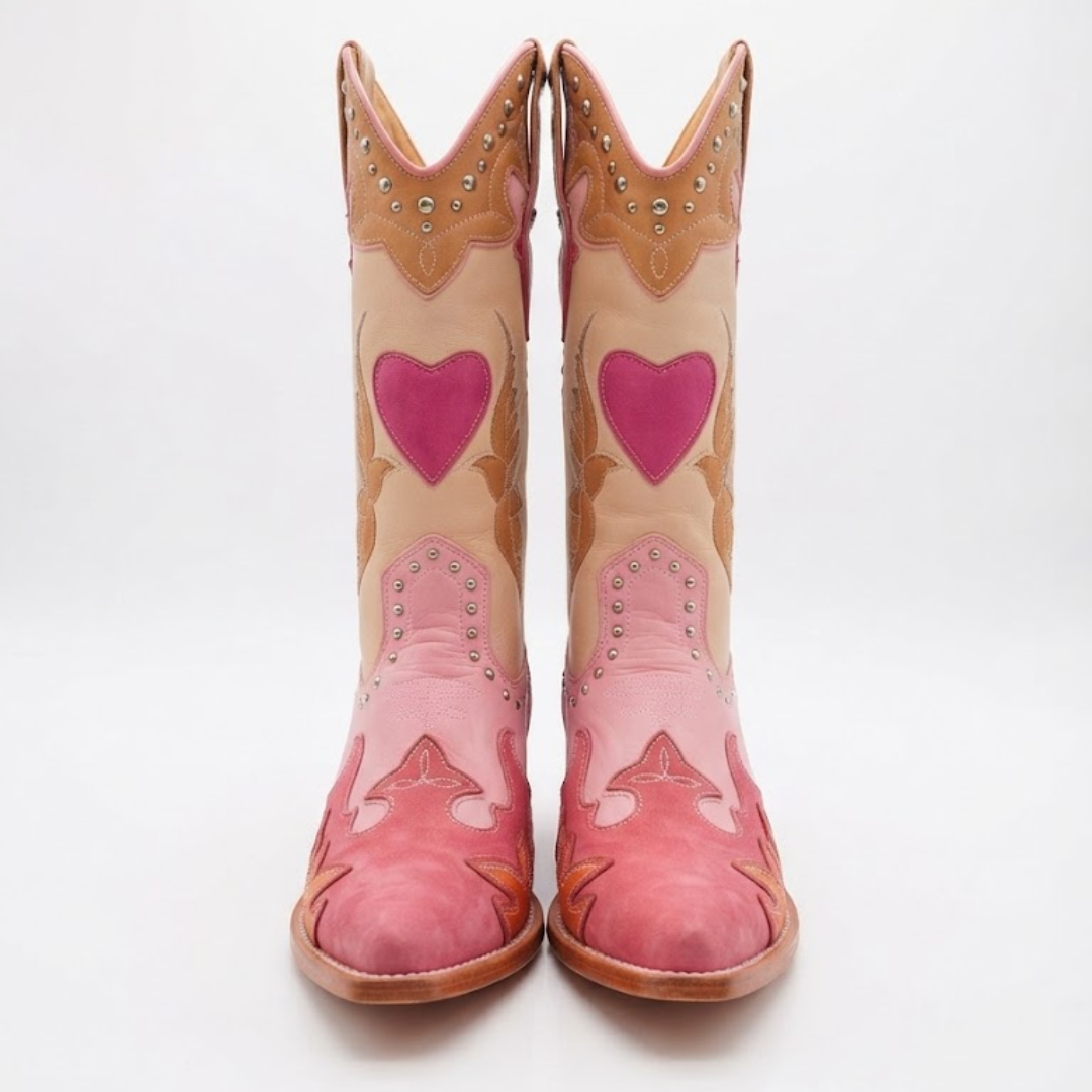 A pair of western cowboy boots in shades of pink and tan, featuring pink heart-shaped patches on the shafts and studded leather details, shown against a plain white background.