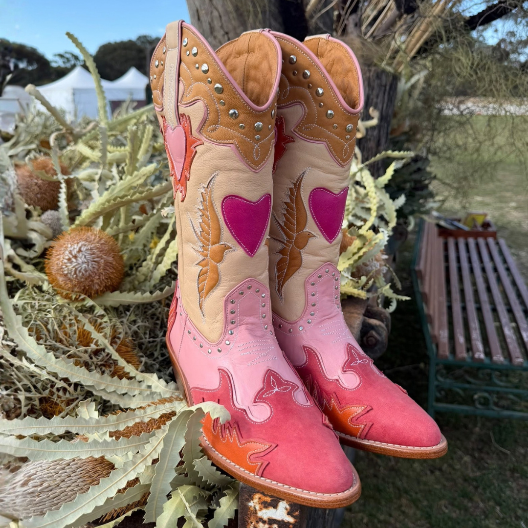 A pair of tall western cowboy boots in pink, peach, and tan suede with heart and feather embroidery, displayed outdoors among dried plants and grass.