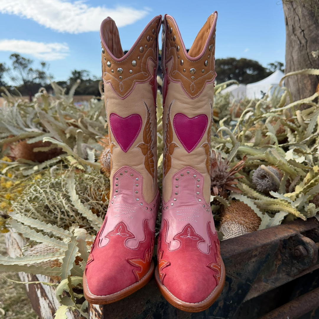 A pair of handcrafted western cowboy boots in pink and tan leather, featuring a bright pink heart on the shafts and a pink gradient foot, displayed outdoors on a rustic wooden crate surrounded by dried plants.