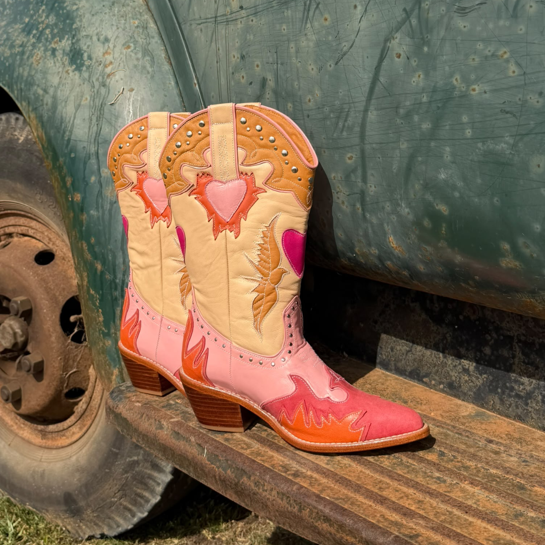 A pair of western cowboy boots with pink, peach, and orange heart and flame designs, leaning against the wooden step of a weathered green truck next to a tire.