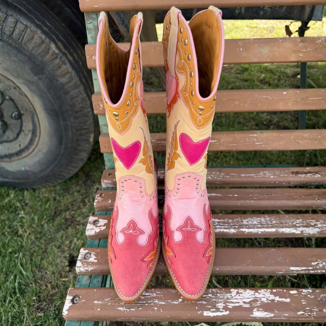 A pair of cowboy boots with pink, orange, and yellow western-style embroidery, displayed on a weathered wooden bench outdoors.