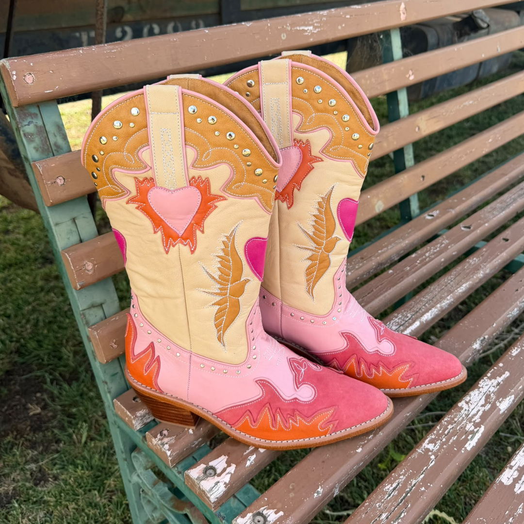A pair of decorative cowboy boots with pink, orange, and beige western designs, placed on a weathered wooden bench outdoors.