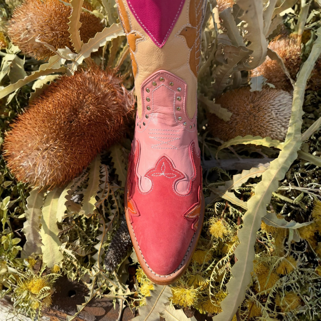 A single western-style cowboy boot photographed from above, placed among dried brown flowers and green leaves outdoors. The boot is primarily pink and tan with decorative stitching and metal eyelets.