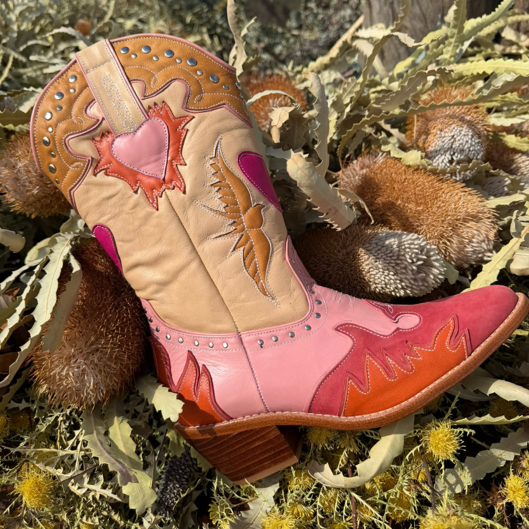 A western-style cowboy boot with pink, peach, and tan leather panels, studded details, and embroidered floral motifs, photographed among dried leaves and brown plant material outdoors.