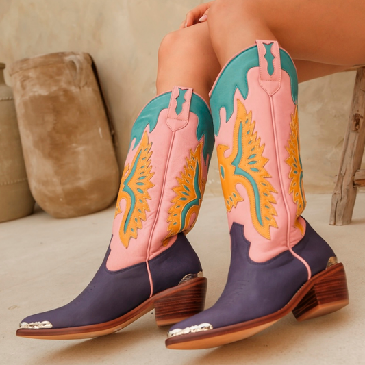 A pair of western cowboy boots worn on a model's feet, featuring a dark purple lower shaft, a pink upper shaft, and teal and yellow embroidered details. The boots have silver conchos on the toes and stacked brown heels.