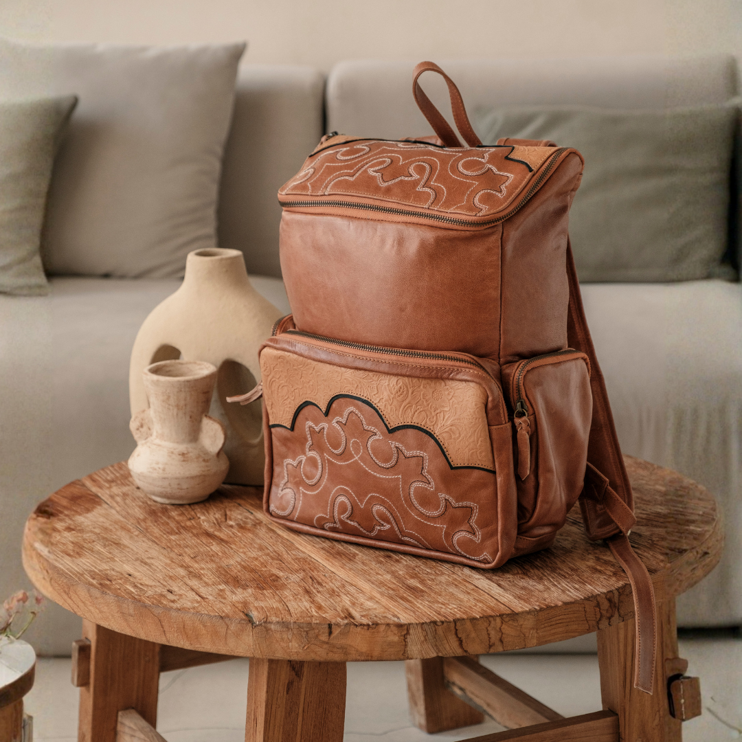 Ridgeback Leather Backpack from House of Hide in brown leather with hand-tooled floral details being displayed on rustic wooden table in cozy living room with ceramic vases