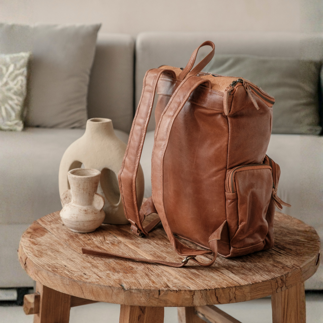 Ridgeback Leather Backpack from House of Hide in brown leather being displayed on round wooden table next to ceramic vases with light gray sofa in background