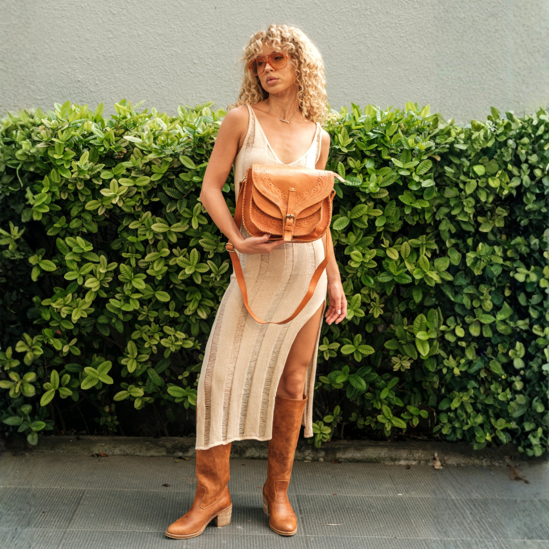 Saddler Crossbody Bag from House of Hide in tan leather being carried by model wearing tan knee-high boots outdoors in front of green hedge