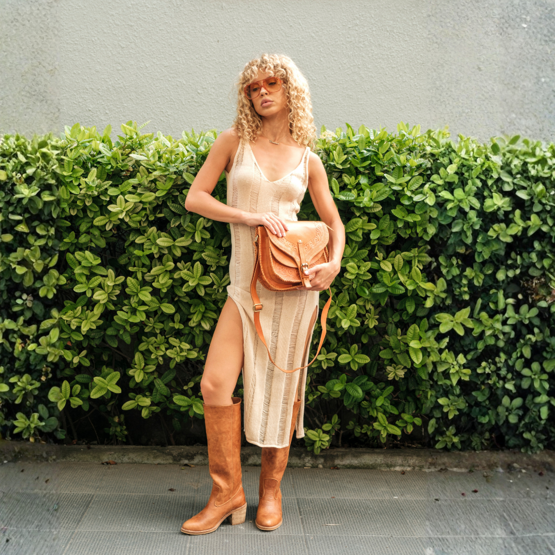 Saddler Crossbody Bag from House of Hide in brown leather being worn by model wearing tan knee-high boots outdoors in front of green hedge
