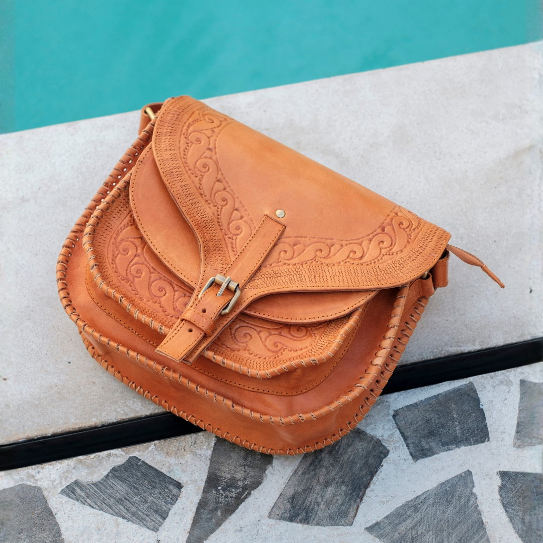 Saddler Crossbody Bag from House of Hide in tan leather with embossed floral detailing on flap and silver-tone buckle being displayed on stone surface with turquoise background